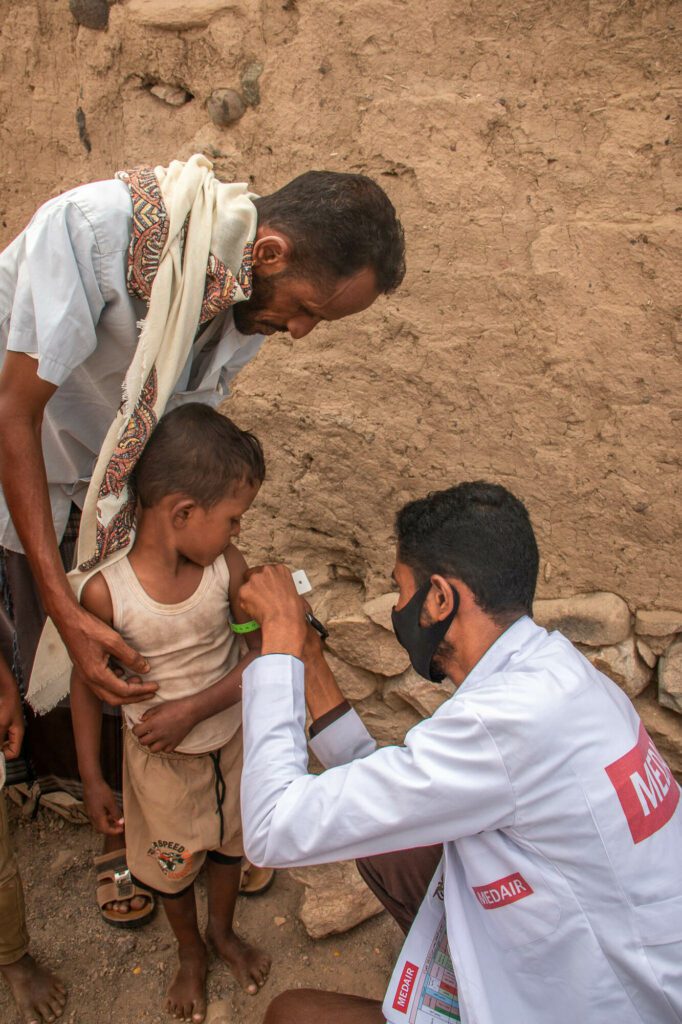 A Medair medical assistant measures the arm circumference of a young boy who stands supported by his father.