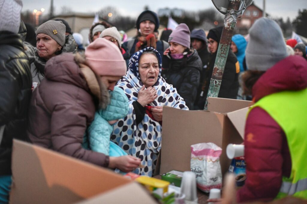 an elderly woman in a shawl stands at a crowded refugee reception point in Poland
