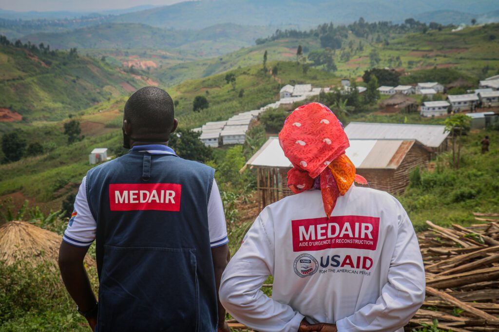 A man and a woman, members of Medair staff, wearing the organization logo on their backs, look out over a valley in DR Congo