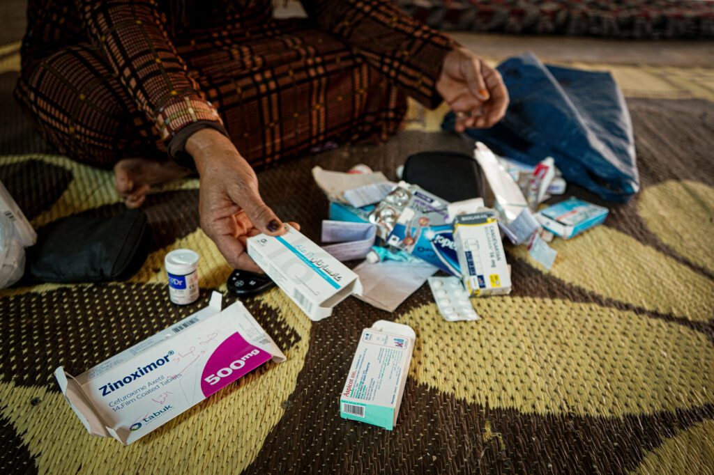 Several empty prescription medications rest on a floor in a tented home whilst a female community member holds an empty box of medication up