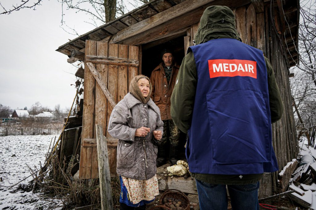 An elderly woman stands in front of her home in Ukraine and talks with a humanitarian. 