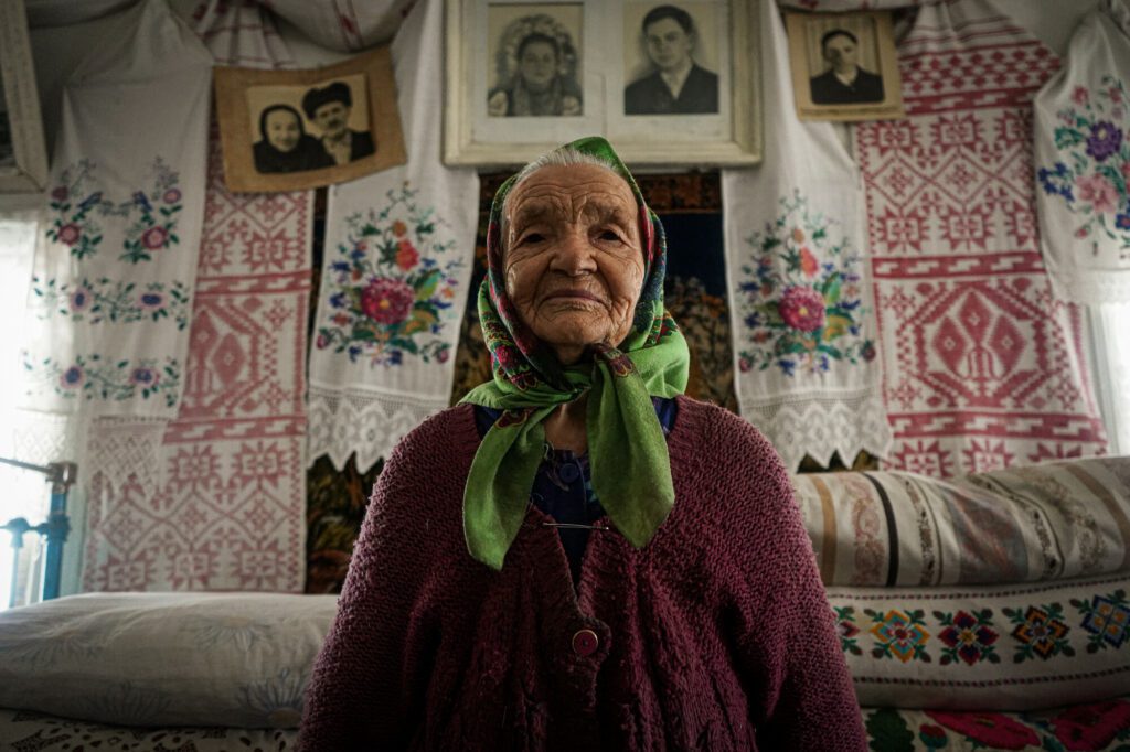 An elderly woman sits on a bed in a room covered with patterned curtains. 