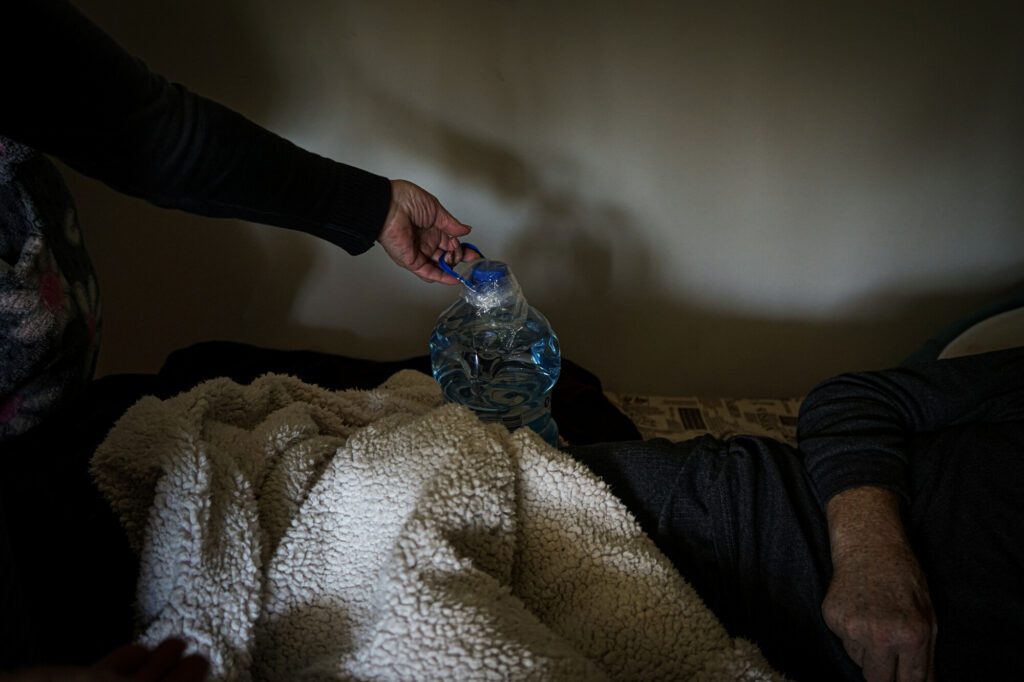 An elderly woman holds a bottle of warm water placed on a bed under sheets.