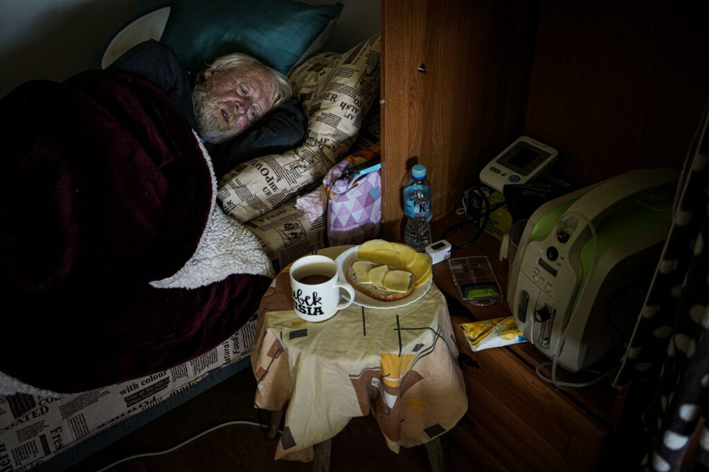  An elderly man sleeps on a bed next to a table with some food on it.