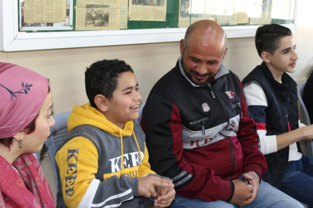 Ein zehnjähriger Gehörloser sitzt lächelnd neben seinem Vater, mit dem er gemeinsam einen Gebärdensprachkurs von Medair in Amman besucht. 