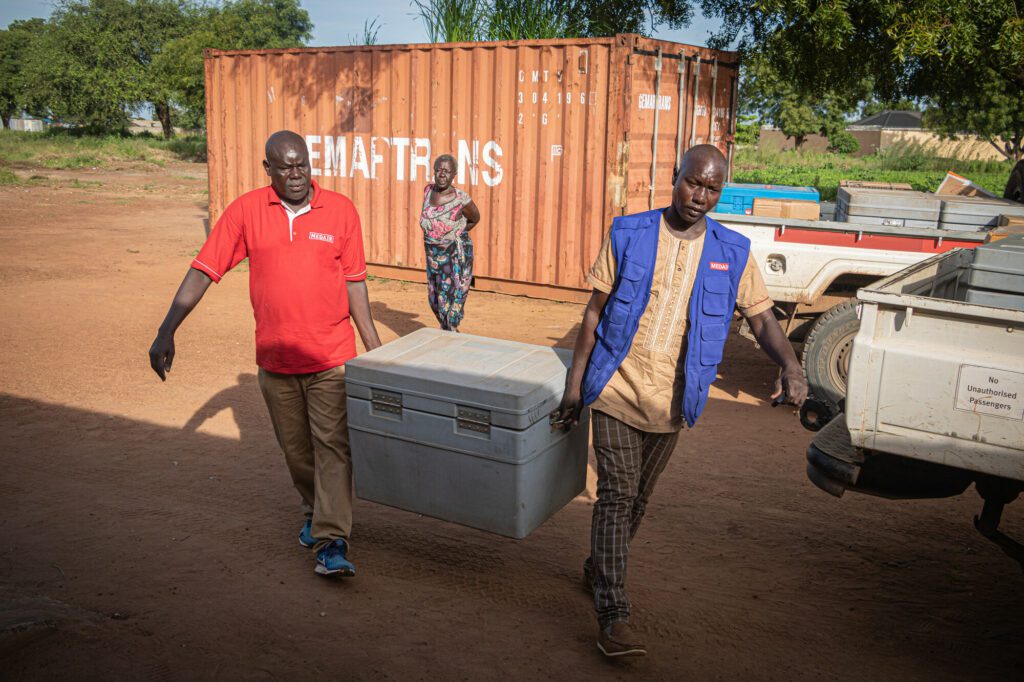 Two male humanitarian aid workers carrying a cooling box with measles vaccines in South Sudan.