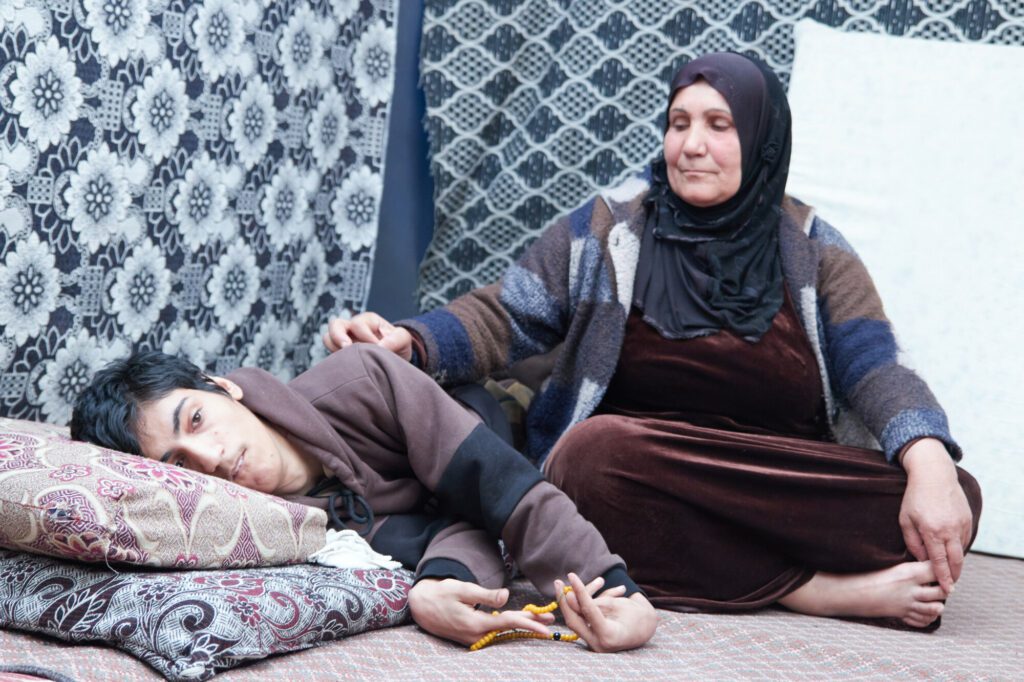 A mother sits beside her son, who is lying down. 