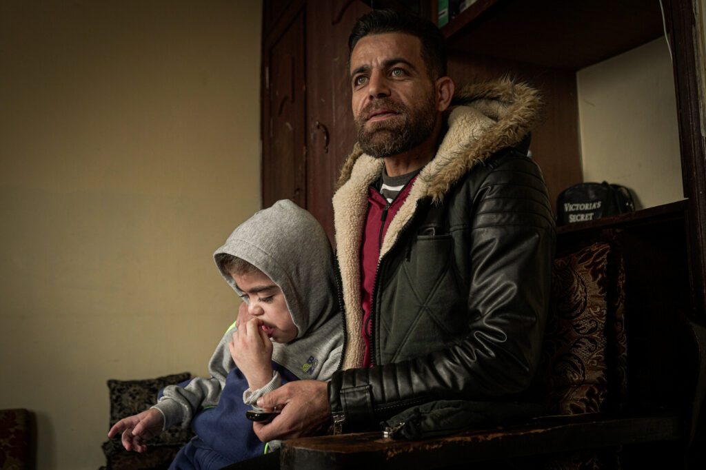Ein syrisches Kind mit Down-Syndrom sitzt im Arm seines Vaters auf einem Stuhl. 