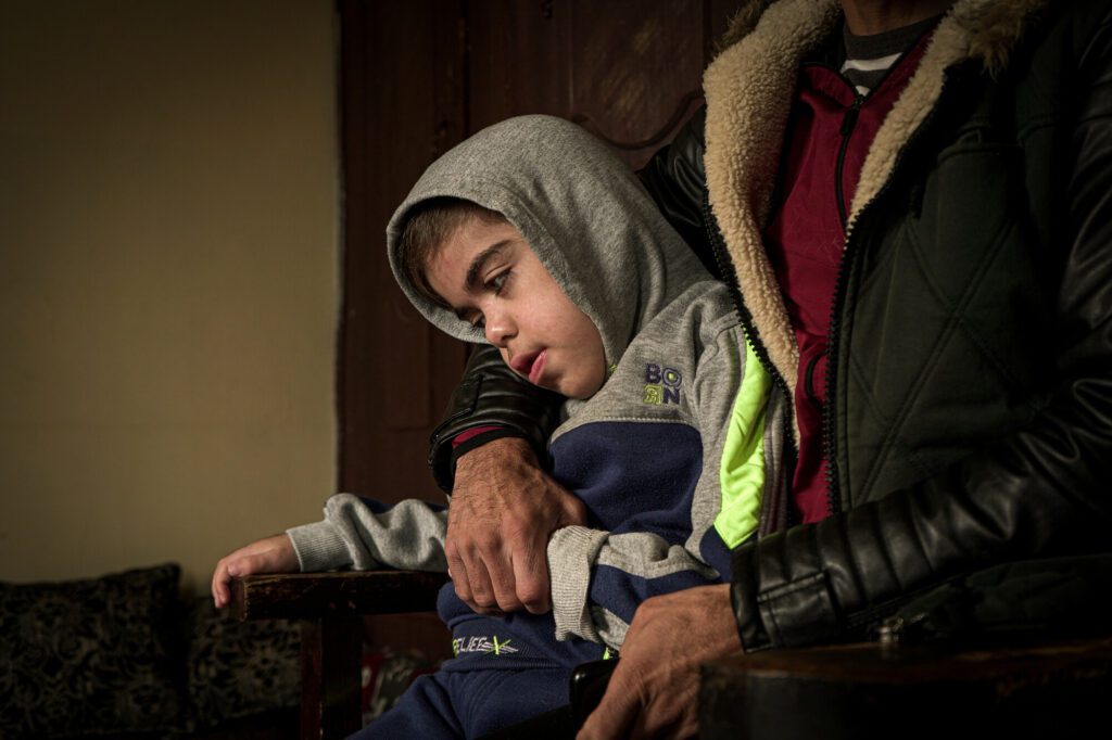 Ein syrisches Kind mit Down-Syndrom sitzt im Arm seines Vaters auf einem Stuhl. 