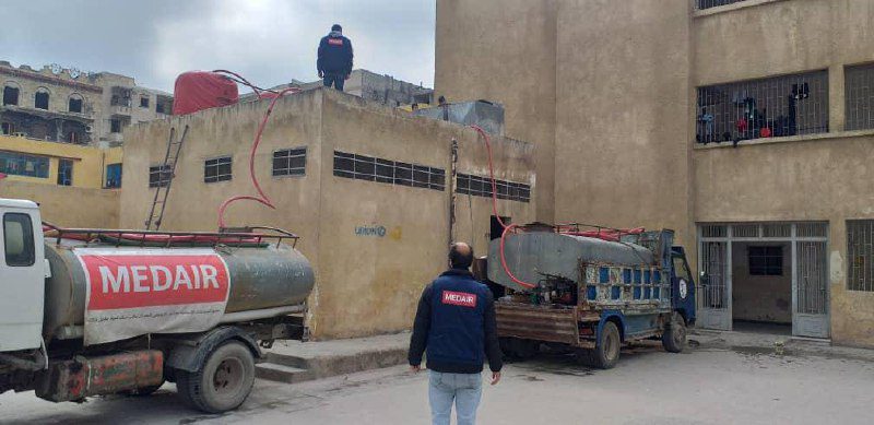 Khaled, employé de Medair, se tient sur le toit de l'école Al Takadum Al Arabi, à Alep, et attend que les réservoirs d'eau soient pleins pour demander à son collègue Yehia, à côté du camion citerne, de fermer le robinet d'eau.