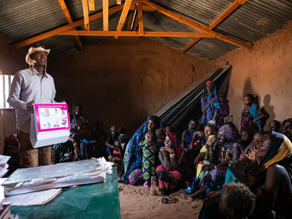 A male humanitarian aid worker shows a Maternal Infant and Young Child Nutrition counselling card to women and children in North Horr, Northern Kenya.