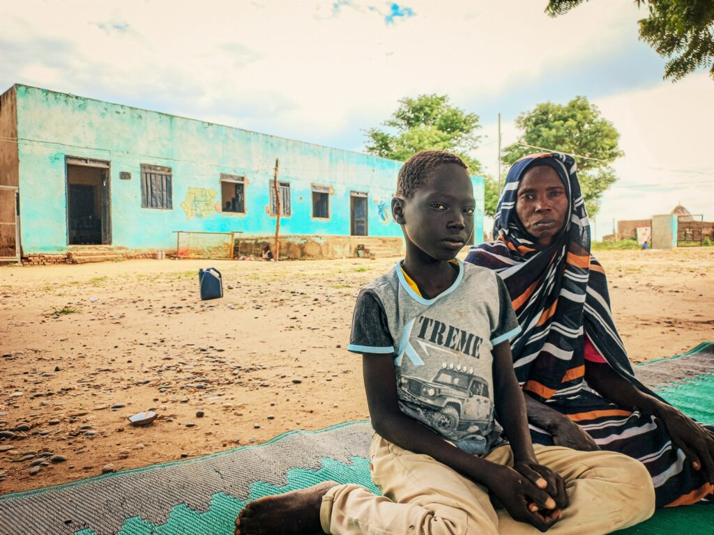 A woman and a boy sitting on a carpet in front of a health facility in Blue Nile, Sudan
