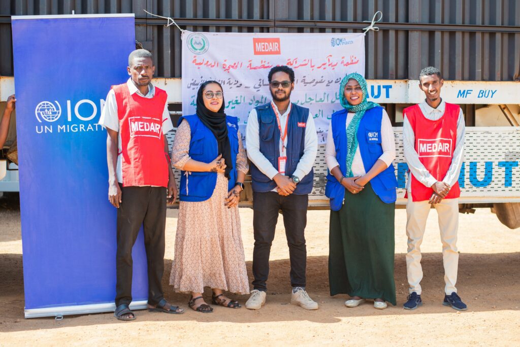 Drie mannelijke en twee vrouwelijke noodhulpmedewerkers staan voor informatieborden tijdens een distributie van hulpgoederen in de staat White Nile in Soedan.